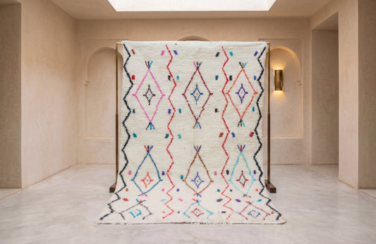 A large Moroccan-style woven rug with a cream base and colorful geometric diamond patterns, displayed on a wooden stand in a minimalist beige interior.