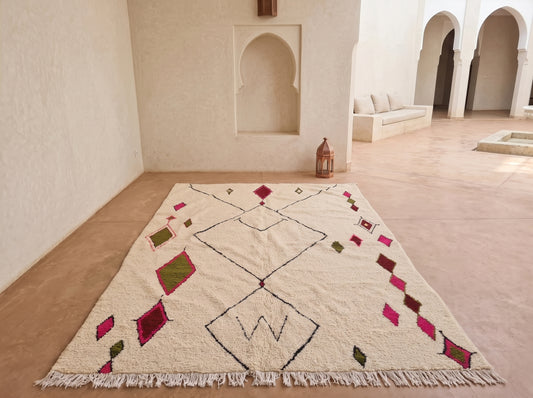 A large Moroccan-style wool rug with a cream base and geometric diamond patterns in black, pink, and olive green, displayed on a terracotta floor in a minimalist Moroccan courtyard.