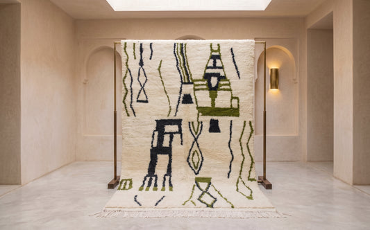 A large woven area rug displayed on a wooden stand in a minimalist beige interior space. The rug features an off-white base with abstract geometric and tribal-inspired patterns in olive green and black.