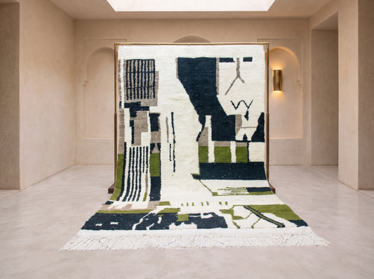A large abstract area rug with a geometric pattern in cream, black, olive green, and beige, displayed hanging from a wooden frame in a minimalist beige gallery space.