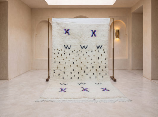 A large white Moroccan wool rug displayed on a wooden stand in a minimalist beige gallery space. The rug features geometric patterns including purple 'X' shapes, purple 'W' shapes, and rows of small black embroidered dots.