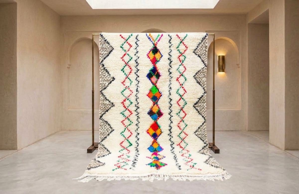 A large woven rug displayed on a wooden stand in a minimalist beige gallery space. The rug features a cream-colored base with black geometric zig-zag patterns and a vibrant, multicolored central diamond motif.