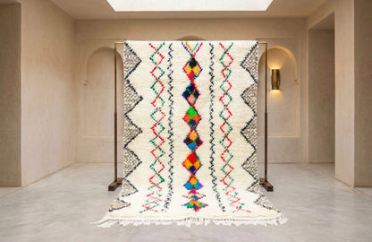 A large woven rug displayed on a wooden stand in a minimalist beige gallery space. The rug features a cream-colored base with black geometric zig-zag patterns and a vibrant, multicolored central diamond motif.