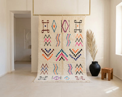 A large woven wall hanging or tapestry with cream-colored fabric and colorful geometric patterns, displayed in a bright, modern minimalist room next to a black vase with dried palm leaves.