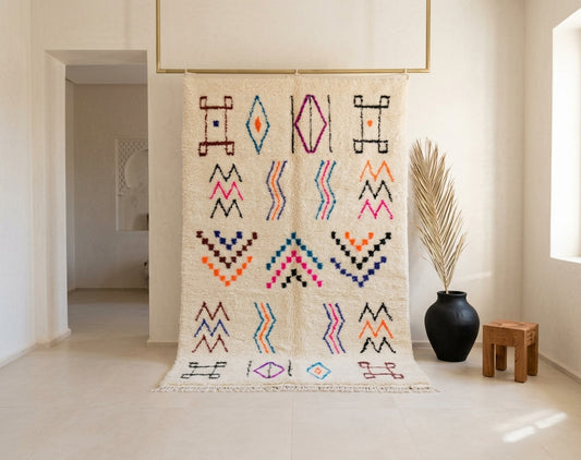 A large woven wall hanging or tapestry with cream-colored fabric and colorful geometric patterns, displayed in a bright, modern minimalist room next to a black vase with dried palm leaves.