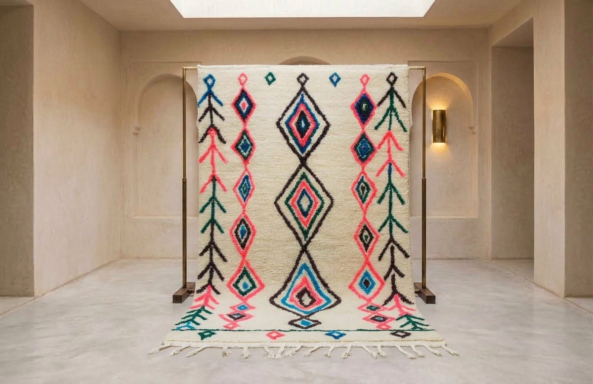 A large woven area rug with a cream base and bold geometric diamond patterns in black, pink, teal, and blue, displayed on a wooden stand in a minimalist beige room.
