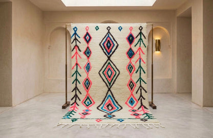 A large woven area rug with a cream base and bold geometric diamond patterns in black, pink, teal, and blue, displayed on a wooden stand in a minimalist beige room.