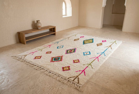 A rectangular cream-colored Moroccan-style rug with colorful geometric diamond patterns and fringe, displayed on a light stone floor in a minimalist room with a wooden bench.