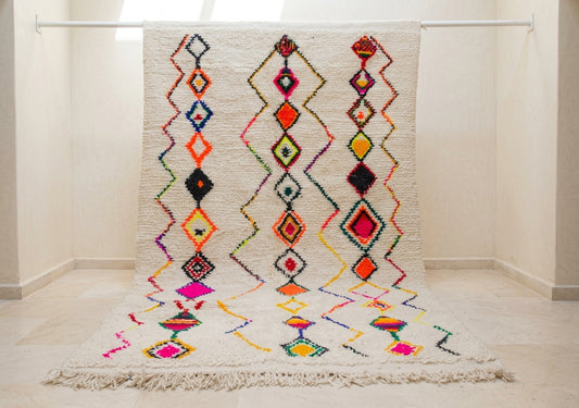 A cream-colored Moroccan-style woven rug with vibrant multicolor geometric diamond patterns, hanging from a white metal rod in a minimalist room with light beige walls and floor.