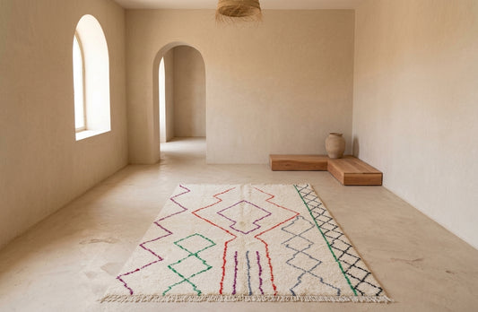 A rectangular area rug with a cream base and colorful geometric patterns, displayed on a light stone floor in a minimalist, warm-toned room with arched windows and wooden benches.