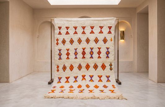 A large woven Moroccan-style rug displayed on a black metal stand in a minimalist beige room. The rug features a cream-colored base with rows of red, orange, and purple geometric diamond patterns and a fringed edge.