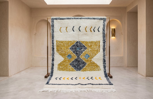 A large Moroccan-style wool rug displayed on a wooden stand in a minimalist, warm-toned gallery space. The rug features a cream background with a central geometric diamond motif in navy blue and gold, bordered by decorative arrow patterns.