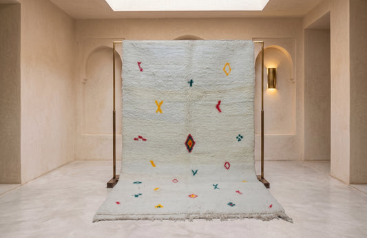A large white Moroccan wool rug with colorful geometric patterns is displayed on a wooden stand in a minimalist, warm-toned interior room with beige walls and arches.