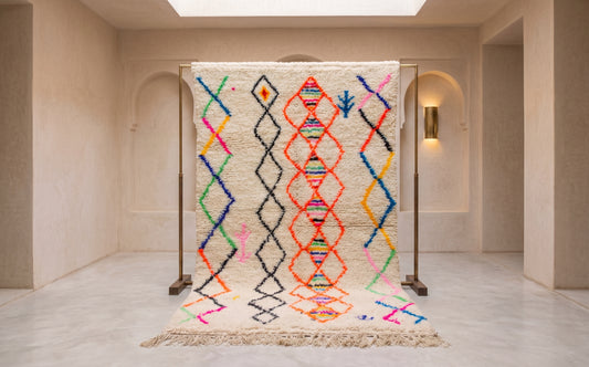 A large woven area rug displayed on a metal stand in a minimalist beige interior space. The rug has a cream base with vertical columns of colorful geometric diamond and zigzag patterns in orange, blue, green, pink, and black, and a fringed bottom edge.
