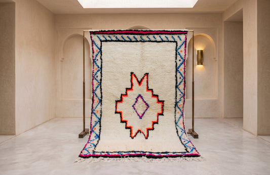 A large woven rug displayed on a metal stand in a minimalist, warm-toned gallery space. The rug features a cream-colored base with a central geometric medallion in red, orange, and purple, surrounded by blue and pink border patterns.
