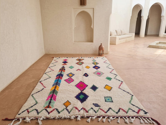 A rectangular Moroccan-style wool rug with a cream base and colorful geometric patterns, displayed on a terracotta-colored floor in a minimalist, sunlit courtyard with arched walls.