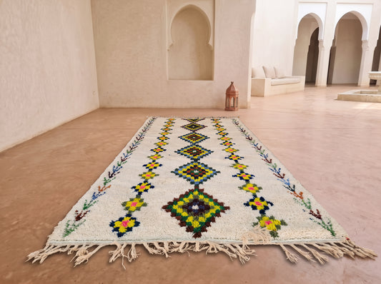 A long Moroccan-style wool rug with a cream base and colorful geometric patterns, laid out on a terracotta-colored floor in a minimalist, sunlit room with arched alcoves.