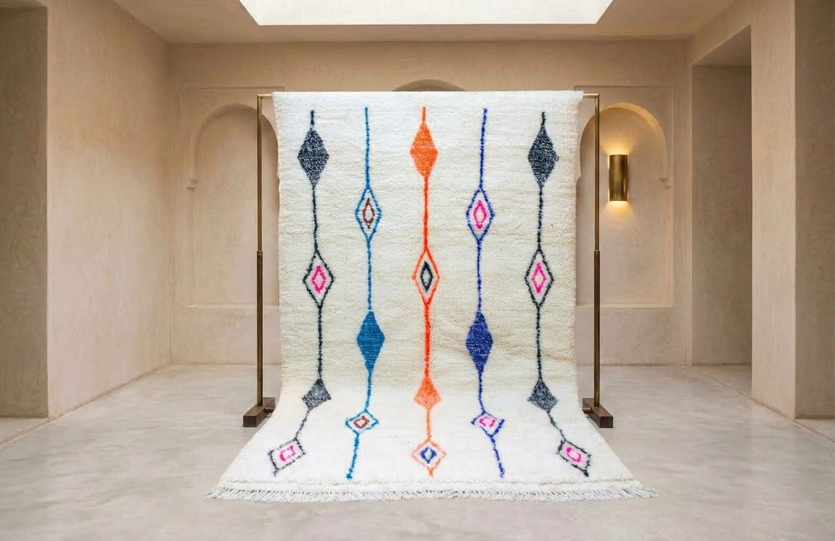A large white Moroccan-style rug with colorful geometric diamond patterns is displayed on a wooden stand inside a minimalist beige gallery space.