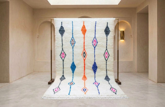 A large white Moroccan-style rug with colorful geometric diamond patterns is displayed on a wooden stand inside a minimalist beige gallery space.