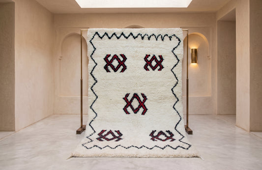 A large white Moroccan-style rug with black zigzag borders and red geometric motifs, displayed on a wooden stand in a minimalist beige interior room.