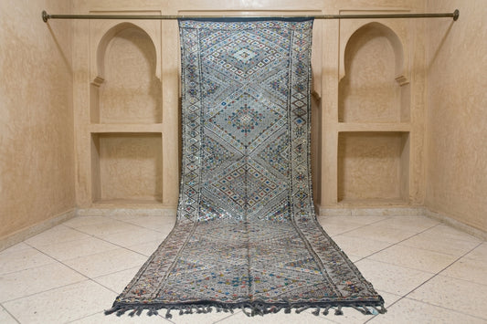 A long patterned rug with fringe is draped over a metal hanging rod inside a beige stone room with arched niches and tiled floor.