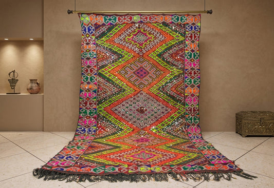 A large, vibrant, multicolored Moroccan-style rug with intricate geometric patterns and black fringe, hanging from a wooden dowel in a warm, neutral-toned gallery room.