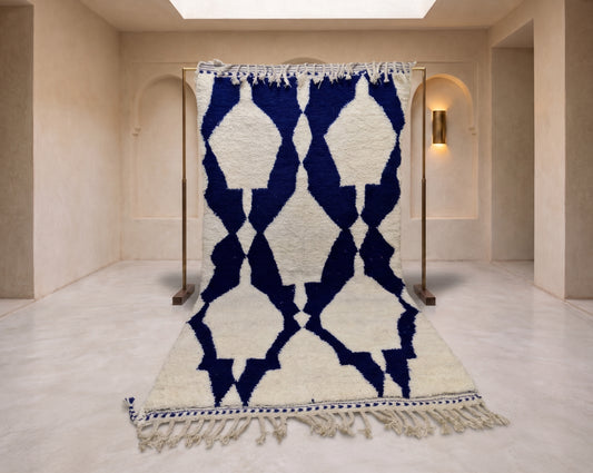 A large woven rug with a bold geometric diamond pattern in deep navy blue and off-white, hanging on a wooden stand in a minimalist beige room with arched walls and a skylight.
