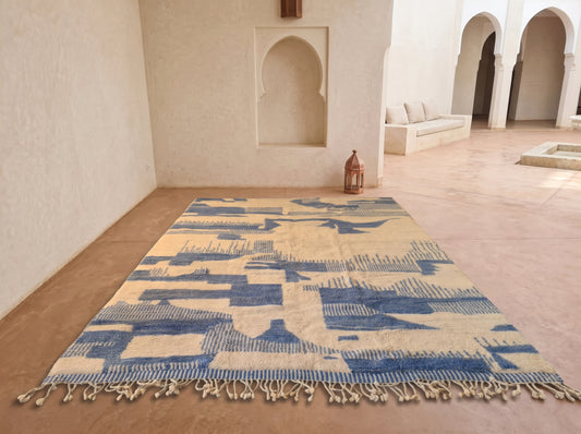A rectangular woven rug with a blue and beige abstract geometric pattern and tasseled edges, displayed on a terracotta floor in a Moroccan-style courtyard with arched alcoves and a bench in the background.