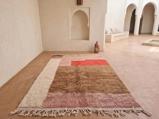 A large Moroccan-style shag rug with geometric color-blocked panels in beige, brown, dusty pink, and terracotta red, laid out on a terracotta floor in a minimalist courtyard with white stucco walls and arches.