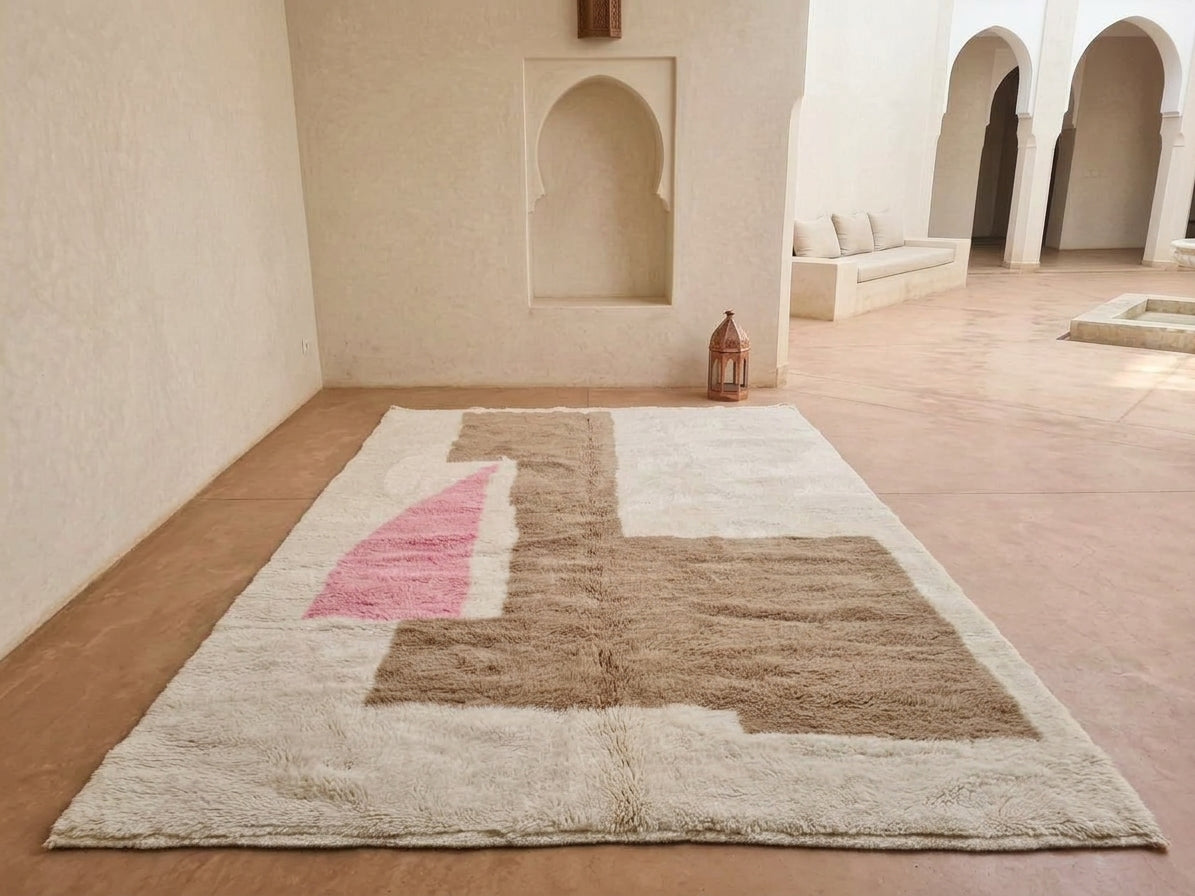 A large rectangular shag rug with an abstract geometric pattern in beige, brown, and pink, displayed on a terracotta floor in a Moroccan-style courtyard with arched alcoves.