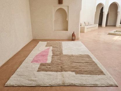 A large rectangular shag rug with an abstract geometric pattern in beige, brown, and pink, displayed on a terracotta floor in a Moroccan-style courtyard with arched alcoves.