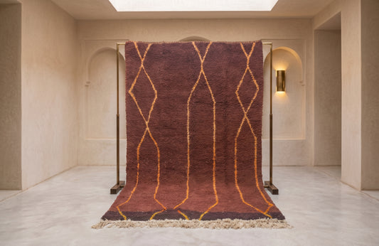 A large Moroccan-style wool rug with a deep reddish-brown base and golden-yellow geometric diamond patterns, hanging on a black metal frame in a minimalist beige room.