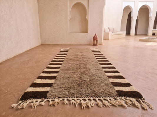 A long rectangular rug with a shaggy taupe center and dark brown and cream striped borders, laid out on a terracotta floor in a Moroccan-style room with arched alcoves.