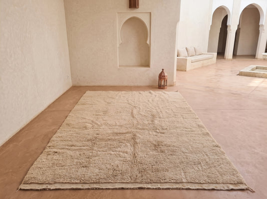 A large rectangular beige rug laid out on a terracotta-colored floor in a minimalist Moroccan-style room with arched alcoves and a small lantern.