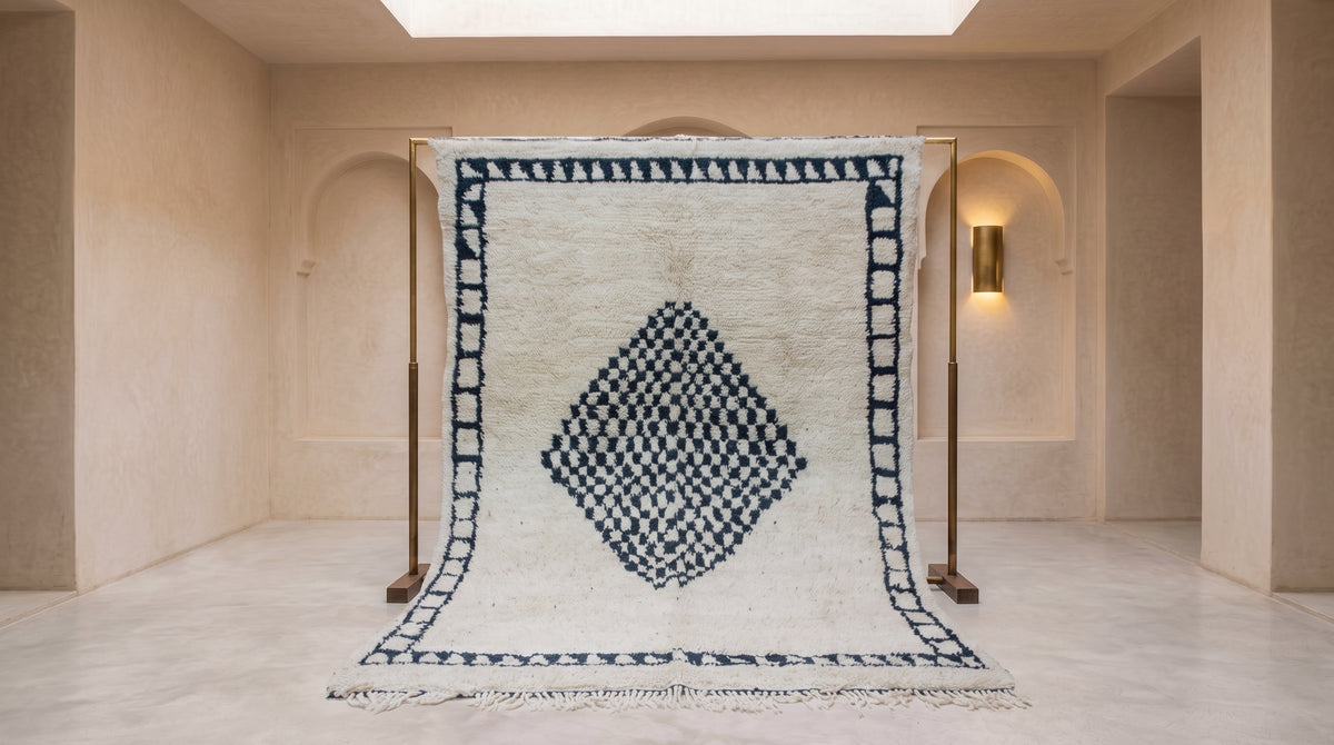A large Moroccan-style wool rug with a cream background and a dark blue geometric diamond pattern, displayed on a wooden stand in a minimalist, warm-toned room.