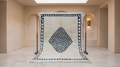 A large Moroccan-style wool rug with a cream background and a dark blue geometric diamond pattern, displayed on a wooden stand in a minimalist, warm-toned room.