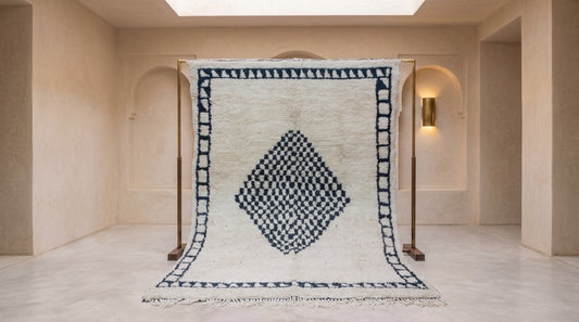 A large Moroccan-style wool rug with a cream background and a dark blue geometric diamond pattern, displayed on a wooden stand in a minimalist, warm-toned room.