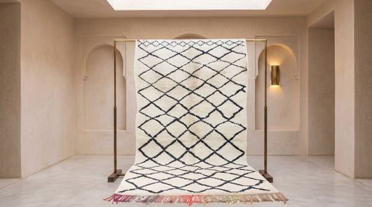 A large Moroccan-style wool rug with a black diamond lattice pattern on an off-white background, hanging on a wooden display stand in a minimalist beige room.