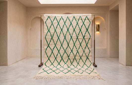 A cream-colored Moroccan-style woven rug with a green diamond lattice pattern, displayed on a wooden stand in a minimalist, warm-toned interior room.