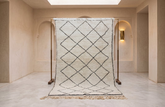 A large rectangular woven rug with a black diamond lattice pattern on a cream background, hanging from a wooden display stand in a minimalist beige interior room.