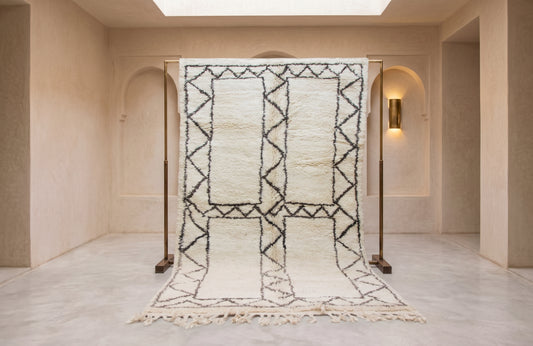 A large Moroccan-style wool rug with a black geometric pattern on an off-white background, hanging from a wooden frame in a minimalist beige hallway.