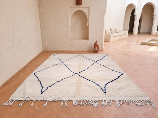 A large off-white Moroccan-style area rug with a dark blue geometric diamond pattern and white fringe, displayed on a terracotta tiled floor in a minimalist courtyard setting.