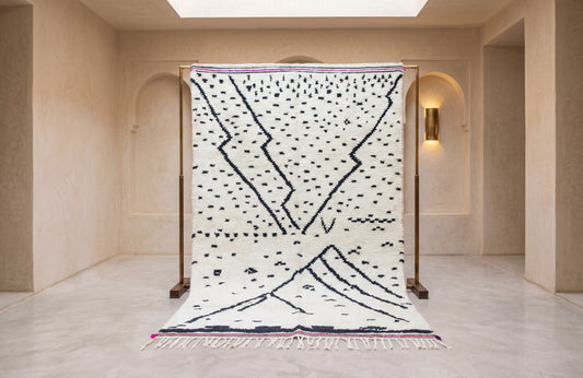 A large white Moroccan-style wool rug with black geometric line patterns and a purple border, displayed on a wooden stand in a minimalist beige room.