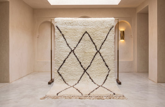 A large off-white shag rug with a black geometric diamond pattern and fringe, displayed on a wooden stand in a minimalist beige room.