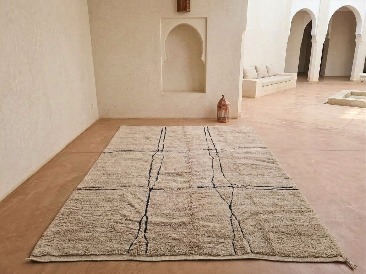 A large rectangular area rug in a warm beige or light brown tone with dark charcoal or black linear geometric patterns, displayed on a terracotta-colored floor inside a minimalist Moroccan-style room with arched alcoves and a bench in the background.