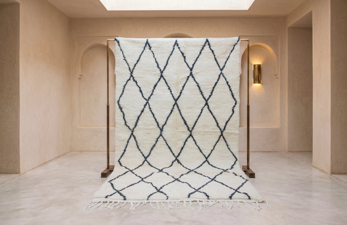 A large white Moroccan-style wool rug with a navy blue geometric diamond pattern, displayed on a wooden stand in a minimalist beige room.