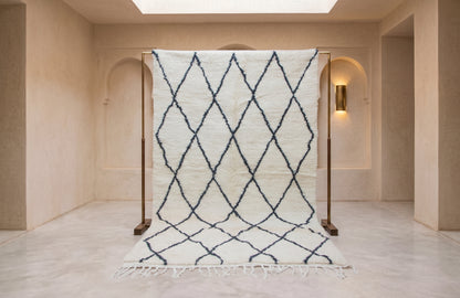 A large white Moroccan-style wool rug with a navy blue geometric diamond pattern, displayed on a wooden stand in a minimalist beige room.