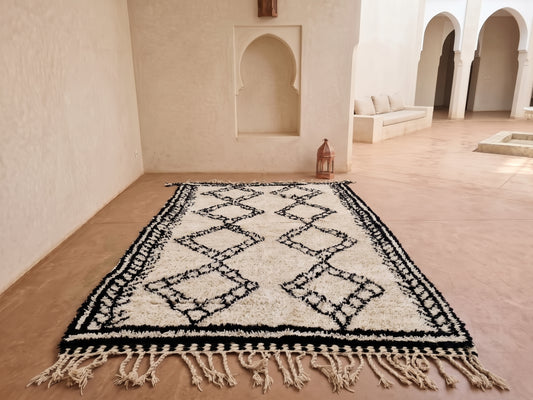 A rectangular woven rug with a black geometric diamond pattern on an off-white background, displayed on a terracotta floor in a minimalist Moroccan-style room with arched alcoves.