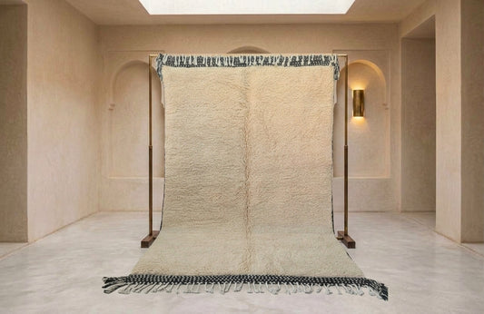 A large cream-colored textured rug displayed on a wooden stand in a minimalist beige room with arched walls and a skylight.