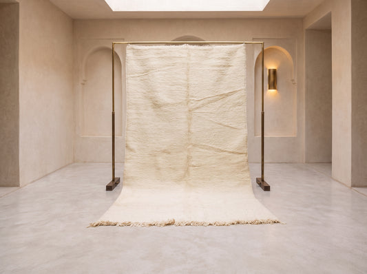 A large cream-colored textured rug draped over a black metal frame stand in a minimalist, warm-toned room with beige walls and a light stone floor.
